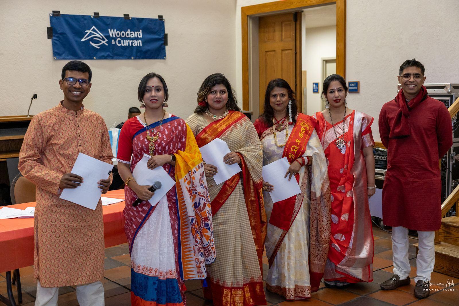 Six organizers in sarees and panjabis holding scripts, sponsor banner behind